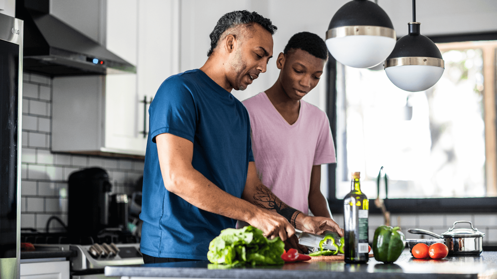 Father and teenage son prepare food in kitchen. Father chops vegetables as son observes.