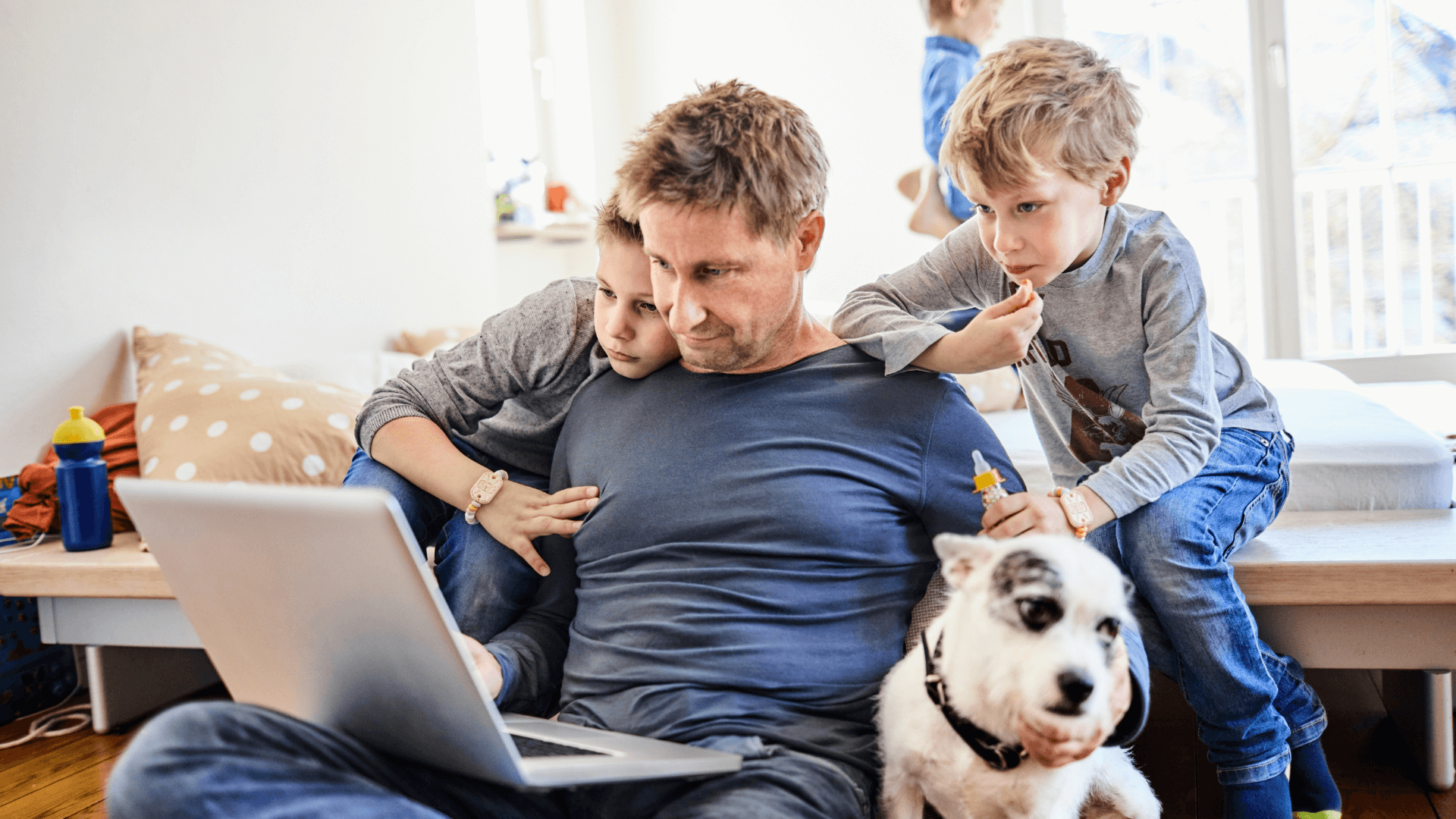 Father sits on floor with laptop on his legs. Two children lean on his shoulders to look at screen.