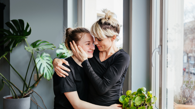 Mother embraces teenage daughter, as they stand next to a window inside their home.